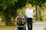 Beautiful woman walking down a lane in park with her little daughter in pushchair, text messaging on mobile phone and smiling. Full length view